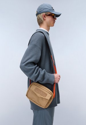 Borsa a tracolla beige con logo in rilievo, tracolla arancione, texture liscia e forma rettangolare. Indossata da una persona con un maglione grigio intrecciato.