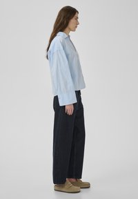 Kvinna med långt brunt hår som bär ljusblå oversized skjorta, vida mörka jeans och beiga loafers, stående i profil mot grå bakgrund.