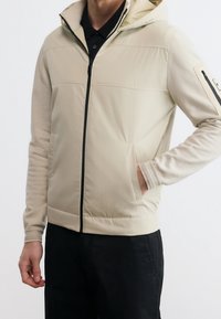 Veste beige zippée avec capuche et fermeture éclair noire, portée sur une chemise noire, une main dans la poche latérale, avec des manches longues jusqu'aux poignets.