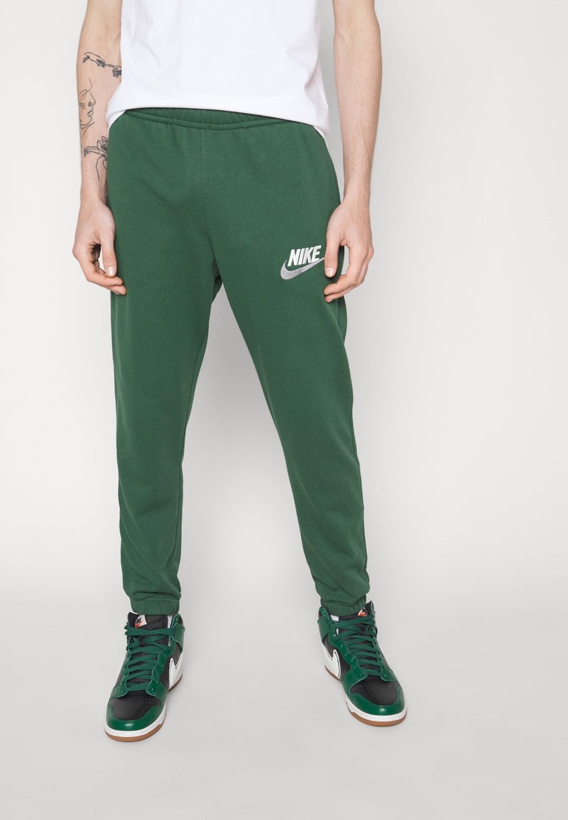 Persona vestida con camiseta blanca, pantalones deportivos Nike verde oscuro y zapatillas altas verdes, negras y blancas, de pie frente a un fondo liso.