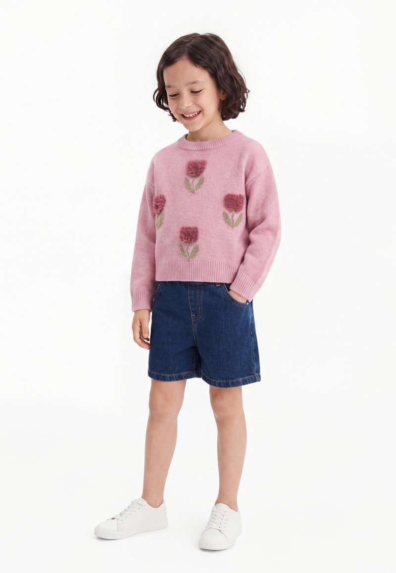 Enfant debout portant un pull rose avec des motifs floraux, un short en jean bleu et des baskets blanches, souriant et regardant vers le bas.