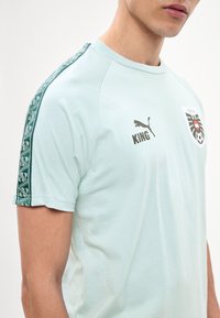 Νέος άνδρας που φοράει ανοιχτό μπλε μπλουζάκι Puma King με σχεδιασμένες πράσινες ρίγες στα μανίκια και ένα έμβλημα ποδοσφαίρου στο στήθος.