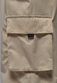 Beige fabric with a textured surface, featuring a large, flat pocket and a black woven label at the center top of the pocket.