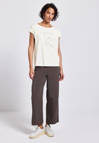 Cremefarvet T-shirt med teksten "INSPIRE" og blomsterdesign, parret med løse, grå-brune bukser og hvide sneakers.
