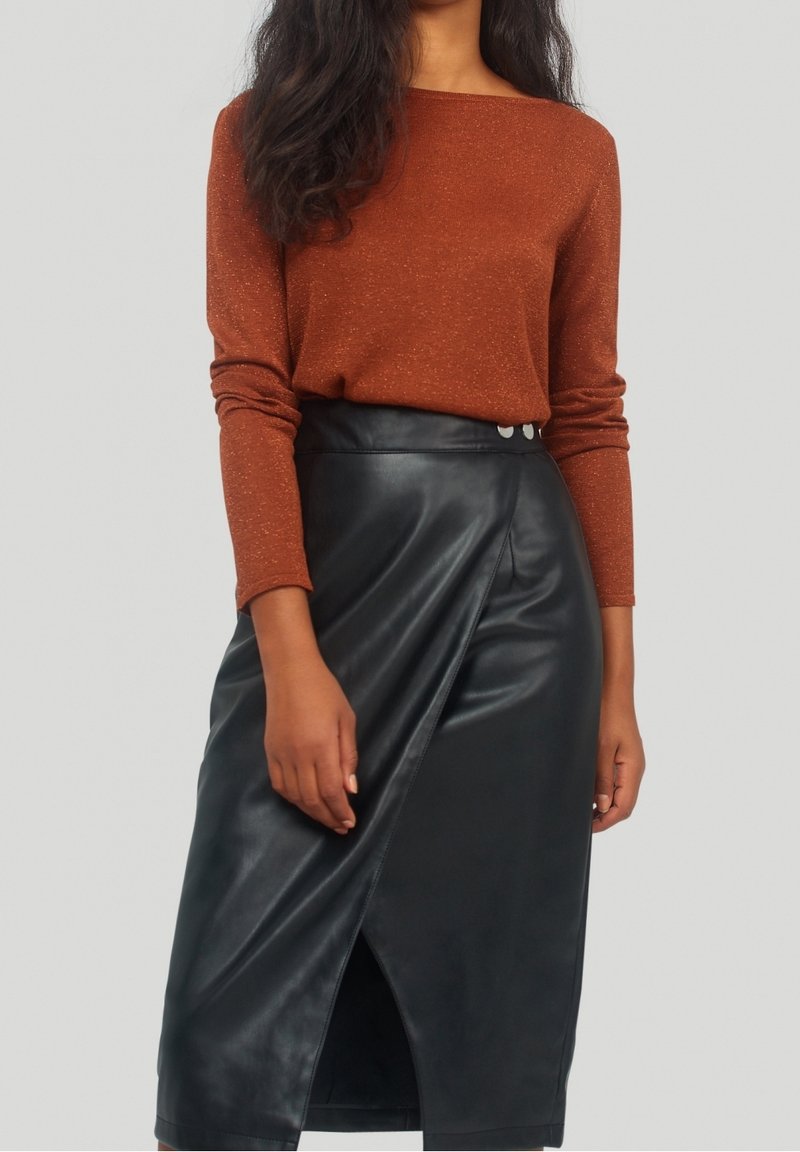 Femme portant un top manches longues couleur rouille rentré dans une jupe portefeuille noire en simili cuir taille haute avec détail de boutons.