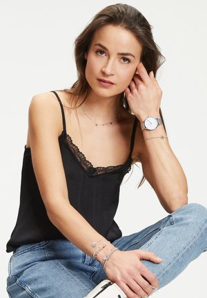Schwarzes Trägershirt mit Spitzenbesatz, Silberne Uhr mit rundem Zifferblatt, übereinander liegende silberne Halsketten, blaue Jeans und gestapelte Armbänder. Neutraler Hintergrund.
