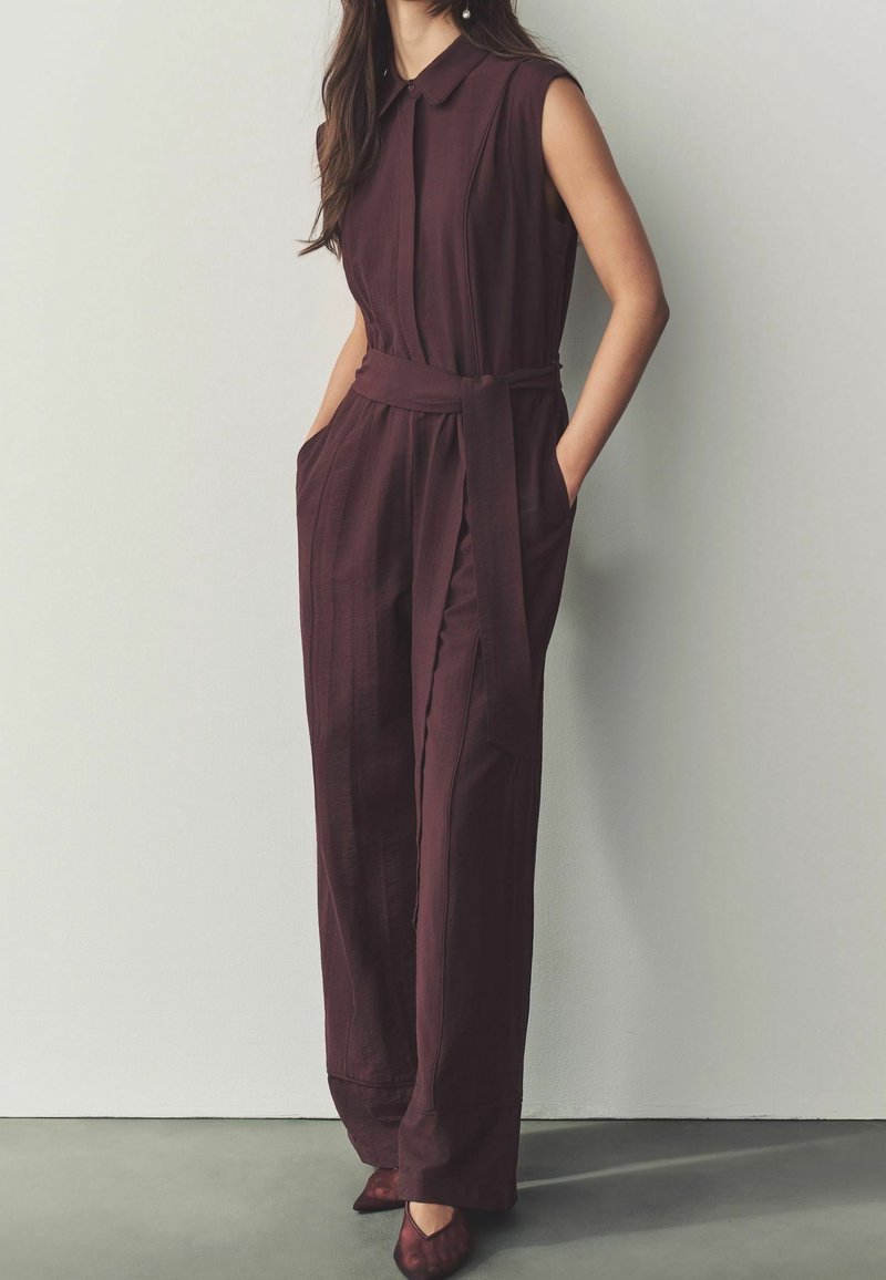 Femme portant une combinaison sans manches violette foncée avec col et taille nouée, debout les mains dans les poches contre un mur clair.