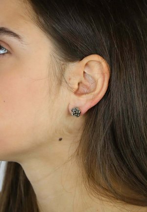 Srebrny kolczyk w kształcie kwiatu na uchu modelki. Kolczyk posiada misternie wykonane detale i gładką teksturę.