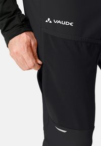 Pantalones elásticos negros con un bolsillo lateral de malla. Logotipo de Vaude en la parte frontal. Textura suave. Presenta mínimos acentos reflectantes.
