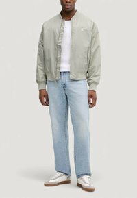 Giubbotto bomber grigio chiaro con zip frontale, polsini a coste e logo semplice. Indossato su una camicia bianca, abbinato a jeans azzurri e sneaker.