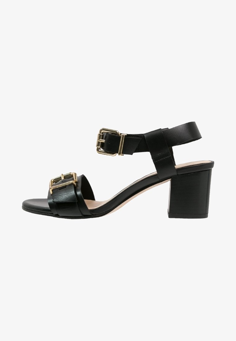 Sandale en cuir noir avec talon bloc, deux brides à boucle dorée — une sur les orteils et une autour de la cheville, design à bout ouvert.