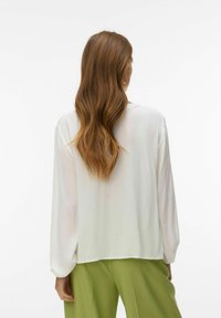 Femme aux longs cheveux bruns ondulés portant un chemisier blanc à manches longues ample et un pantalon vert clair, vue de dos sur un fond uni.