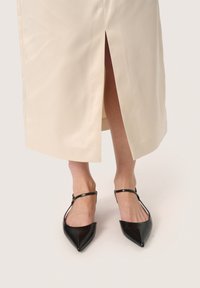 Abito di raso crema con uno spacco laterale, abbinato a tacchi a slingback neri a punta con fasce sottili.