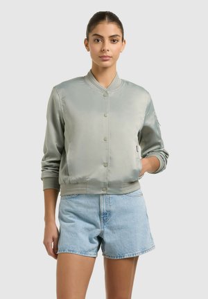 Kvinde stående vendt fremad iført en lysegrå satin bomberjakke og lyseblå denimshorts mod en ensfarvet baggrund.