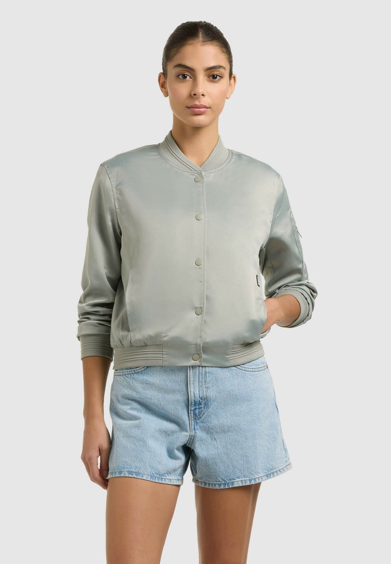 Femme debout, face à l'avant, portant une veste bomber en satin gris clair et un short en denim bleu clair sur un fond uni.