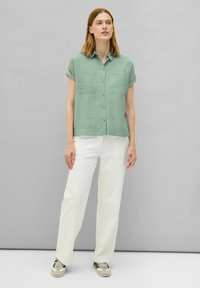 Camicia verde chiaro a maniche corte con bottoni e due taschini sul petto, abbinata a pantaloni bianchi con trama e sneakers beige e verdi.