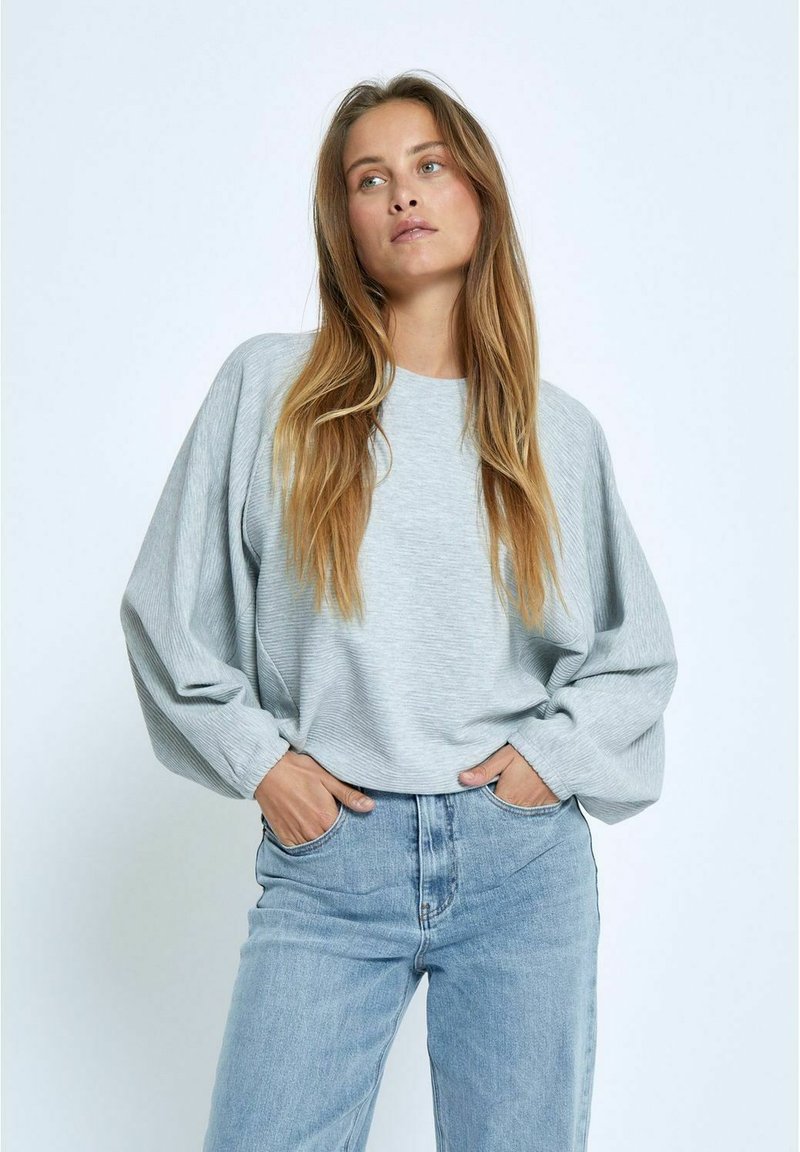 Heller grauer Langarm-Sweatshirt mit strukturiertem Finish, lockerer Passform und überschnittenen Schultern, kombiniert mit hochgeschnittenen blauen Jeans.