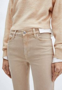 Beige farkut, joissa on viisi taskua, metallinappikiinnitys ja hienovarainen ompelu. Yhdistetty pehmeään, vaaleaväriseen villapaitaan.