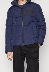 Mörkblå vadderad jacka med dragkedja framtill, featuring en logotyp på bröstet, horisontell quiltning och en huva, tillsammans med svarta byxor.