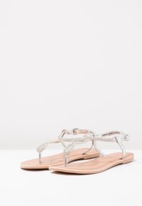 Hopeaketjuiset, tumman hopeansävyiset litteät sandaalit, joissa on yksinkertainen muotoilu. Niissä on varvaslenkki ja nilkkahihna. Pohja on vaaleanbeige ja litteä.