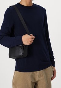 Zwarte leren crossbodytas met ritssluiting, rechthoekige vorm, gladde textuur en verstelbare riem, gedragen over een donkerblauwe trui en beige broek.