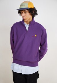 Lilla fleece pullover med kvart-zip, ribbet kant og broderet logo. Laget over en lyseblå og hvid stribet skjorte.