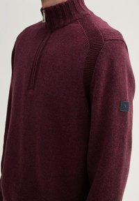 Burgunderfarbener Pullover mit hohem Kragen und halbem Reißverschluss, ausgestattet mit strukturierten Schulterakzenten und einem Logo-Patch an den Ärmeln.