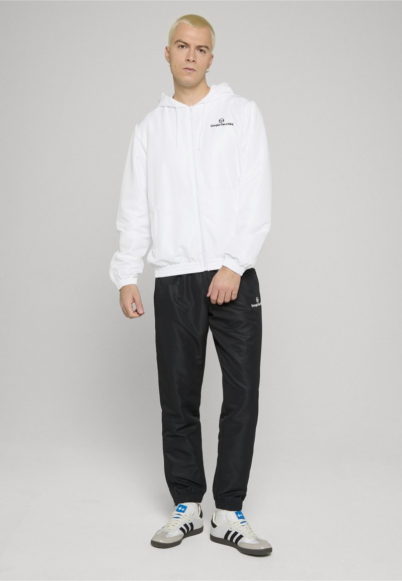 Sudadera blanca con cremallera y puños elásticos, pantalones deportivos negros con logo y zapatillas blancas con rayas negras. Tejido suave y ligero.