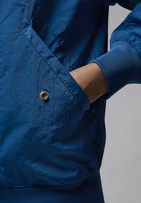 Mano parcialmente dentro del bolsillo de una chaqueta azul con puños acanalados y botón a presión en el bolsillo.