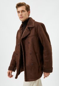 Chaqueta marrón de doble botonadura con un cuello y puños de borrego texturizado, con un material exterior suave y bolsillos laterales.