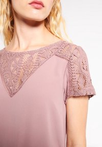 Blouse lavande avec un empiècement en dentelle et des manches courtes. Tissu lisse, texture délicate et accents au crochet aux épaules.