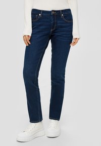 Jeans en denim bleu foncé avec une coupe ajustée, des coutures contrastantes et des poches. Associés à des baskets blanches à semelle épaisse.