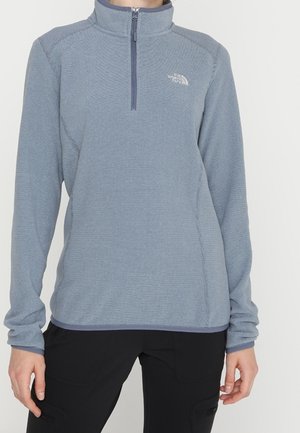 Jasnoniebieska bluza polarowa z suwakiem do ćwiartki i długimi rękawami oraz logo The North Face, noszona przez osobę w czarnych spodniach, stojącą na jednolitym tle.