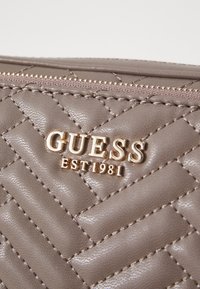 Portafoglio in tessuto sintetico marrone trapuntato con logo dorato in rilievo "GUESS EST 1981" e chiusura a zip lungo il bordo superiore.