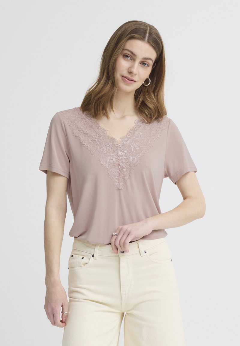 Jeune femme aux cheveux mi-longs portant un haut rose bordé de dentelle et un pantalon taille haute écru, posant avec une main sur la taille.
