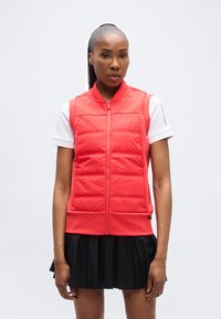 Γυναίκα με σκούρο δέρμα και μαύρη αλογοουρά, που φοράει κόκκινο γιλέκο με ραφές πάνω από λευκό κοντομάνικο πουκάμισο και μαύρη πλισέ φούστα, στέκεται με το πρόσωπο προς τα εμπρός.