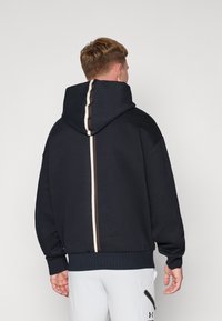 Marineblaues Hoodie mit großer Kapuze, gerippten Bündchen und Saum sowie einem vertikalen Streifendetail in Creme und Braun entlang der Rückenmitte.