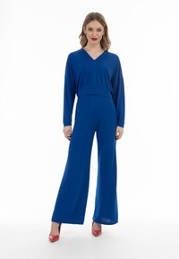 Tuta blu con scollatura a V, maniche lunghe e vita aderente. Pantaloni svasati che toccano il pavimento. Indossata con scarpe rosse con tacco alto. Tessuto liscio.