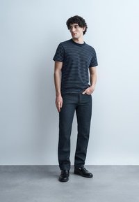 Mladi moški stoji ob svetlem zidu, oblečen v črno-belo črtasto majico, temne kavbojke in črne loafer čevlje, gleda v stran.