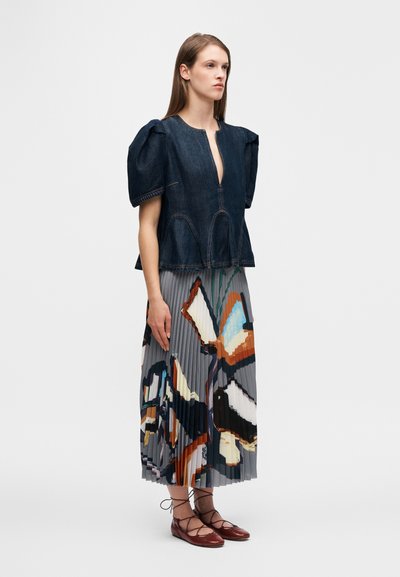 Τοπ από denim με φουσκωτά μανίκια, βαθύ ντεκολτέ σε σχήμα V και ραφτές λεπτομέρειες, συνδυασμένο με πλισέ φούστα με πολύχρωμο αφηρημένο σχέδιο. Καφέ δερμάτινα παπούτσια με κορδόνια.