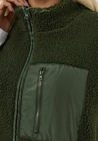 Olivgrön zip-up väst med en texturerad fleecekropp, släta nylonaccenter och en ficka med dragkedja på vänster sida.
