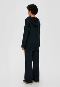 Dunkelgrüner Pullover mit Kapuze, langen Ärmeln und geripptem Saum, kombiniert mit weiten, gestreiften navyblauen Hosen und Sneakers mit kontrastierender Sohle.