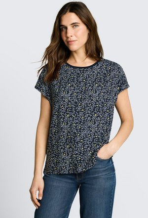 Kobieta o długich brązowych włosach, ubrana w granatową bluzkę na krótkie rękawy w kwiatowy wzór i niebieskie dżinsy, stoi z jedną ręką w kieszeni.