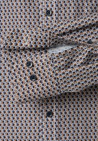 Hemd mit Kragen in geometrischem Muster in Braun, Marineblau und Creme. Besitzt dunkle Knöpfe und einen strukturierten Stoff mit sichtbarem Manschetten.