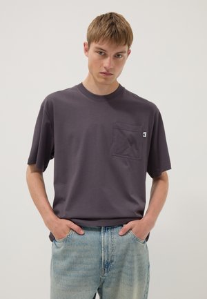 Joven con camiseta gris oscuro con bolsillo y vaqueros azul claro con las manos en los bolsillos, de pie contra un fondo claro y liso.