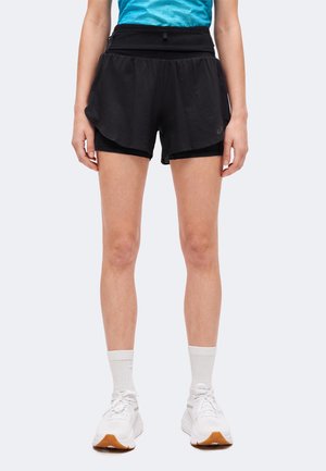 Personne portant un short de sport noir avec une couche de compression intérieure, des chaussettes blanches et des chaussures de course blanches, debout sur un fond uni.