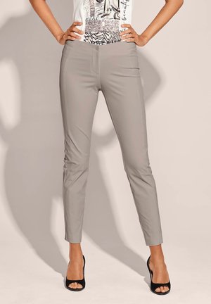 Femme portant un pantalon slim gris clair et des talons ouverts noirs, mains posées sur les hanches, sur fond neutre.