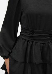 Juoda ilgomis rankovėmis suknelė su surišta juosmens dalimi, sluoksniuotu pepelmu ir lygiu, lengvai krentančiu audiniu.