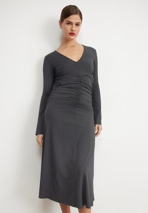 Femme portant une robe midi grise foncée à manches longues et froncée avec un décolleté en V, se tenant devant un mur blanc, faisant face à l'avant.