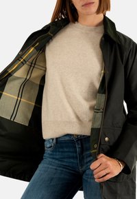 Zwarte buitenjas met een groen geruit voering, voorzien van een rits en knopen. De binnentrui is crèmekleurig, met een geribbelde zoom. De jeans zijn blauw.